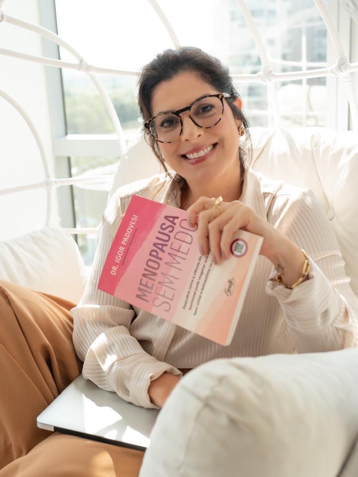 Renata Trivilini sorrindo, sentada em uma poltrona, segurando um livro sobre menopausa